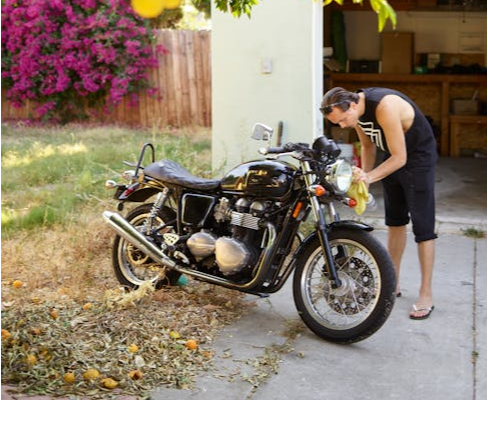 A man wearing a black tank top cleans a motorbike in an outdoor garage space surrounded by greenery.