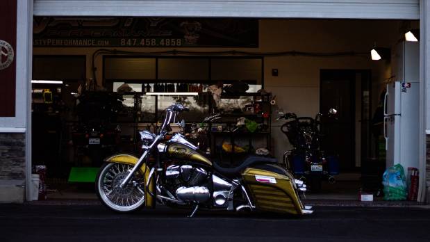 classic home motorcycle, relaxed expression, sitting on seat, photorealistic, vintage garage setting with retro advertisements and old oil drums, highly detailed, polished metal, 24mm lens, sepia tones, morning sunlight streaming in
